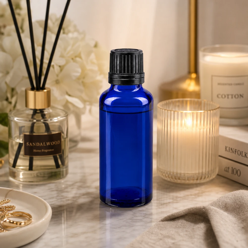 Frasco de cristal azul de 30 ml con tapón cuentagotas negro sobre mármol, junto a una vela perfumada y un ambientador
