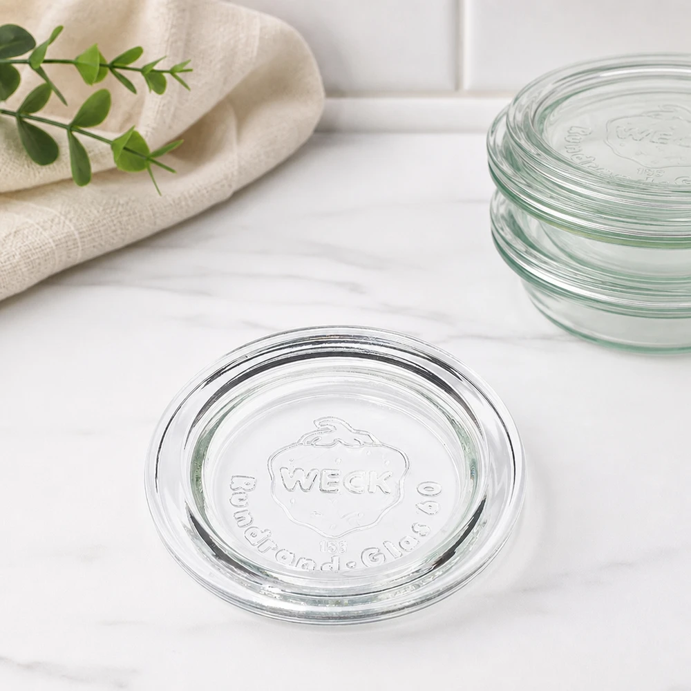 Tapa para tarro WECK RR60 Tapa de cristal WECK RR60 vista apilada de varias tapas superpuestas sobre mármol con paño de cocina y rama verde