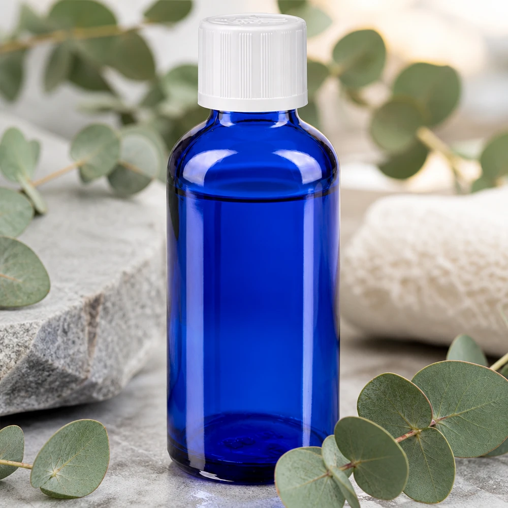 Frasco de farmacia azul cobalto de 50 ml con tapón KiSi blanco sobre una piedra natural entre ramas de eucalipto