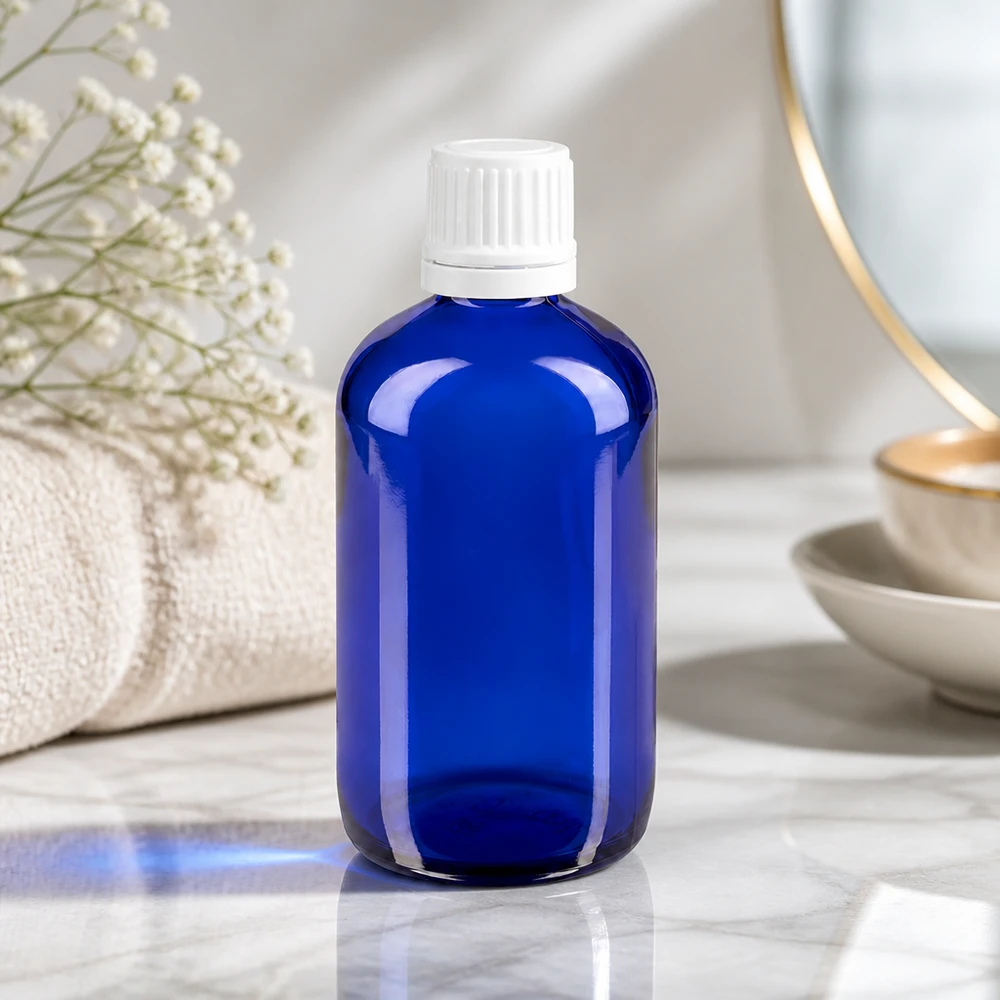 Botella de cristal azul de 100 ml con tapón OV blanco sobre mármol, junto a una toalla y flores secas