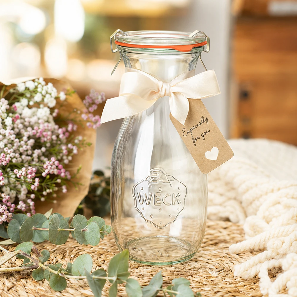 Botella de zumo WECK RR60 de 530 ml decorada con una cinta blanca y una etiqueta de regalo dispuesta entre flores