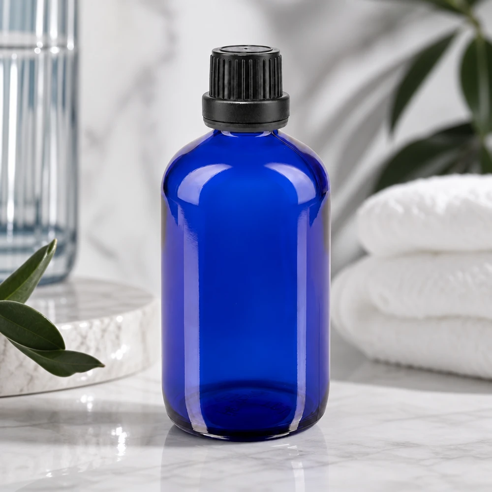 Botella de cristal azul de 100 ml con tapón negro estriado junto a toallas blancas y plantas verdes