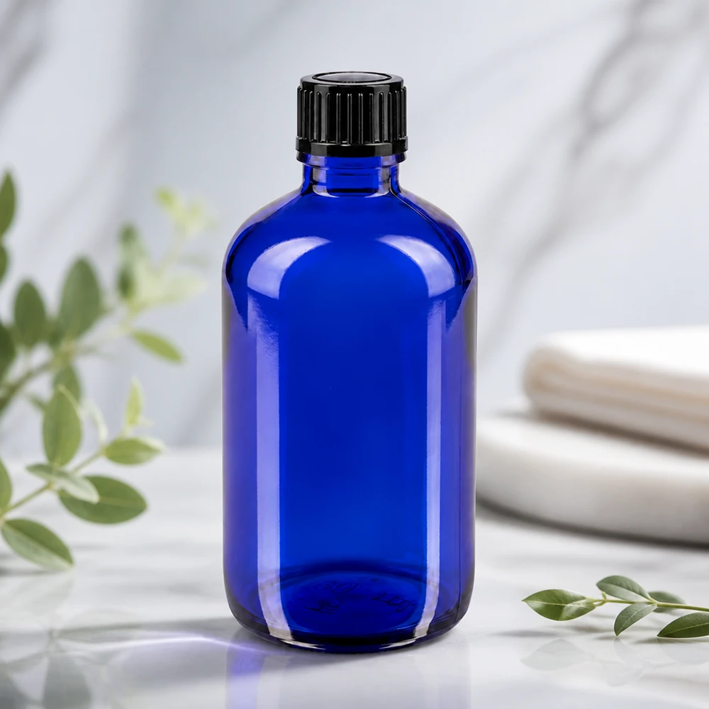 Botella de cristal azul de 100 ml con tapón negro estriado sobre una superficie de mármol blanco con ramitas de eucalipto