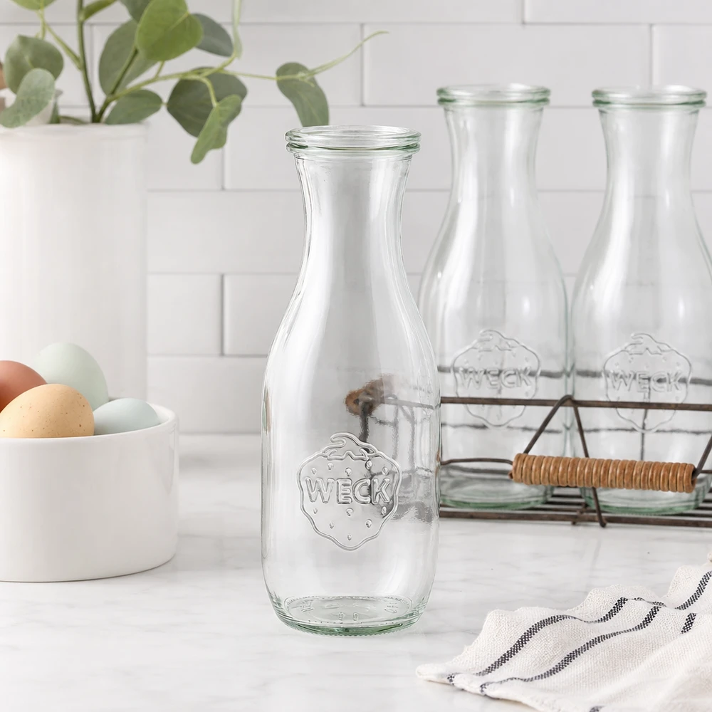 Botella para zumos de 1062 ml con tapa de cristal WECK RR60 WECK botella de zumo 1062ml botella de vidrio vacía en la cocina blanca moderna con cáscara de huevo cesta de alambre y la planta