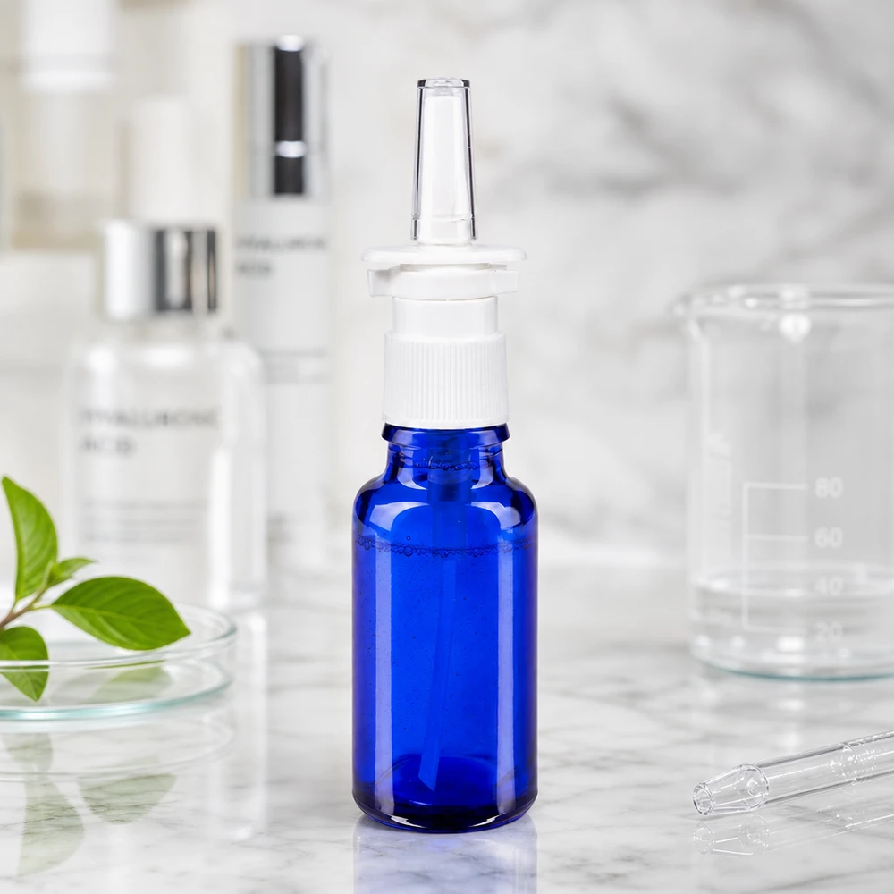 Frasco de spray nasal de 20 ml de cristal azul de farmacia con pulverizador blanco, junto a un vaso de precipitados y ácido hialurónico
