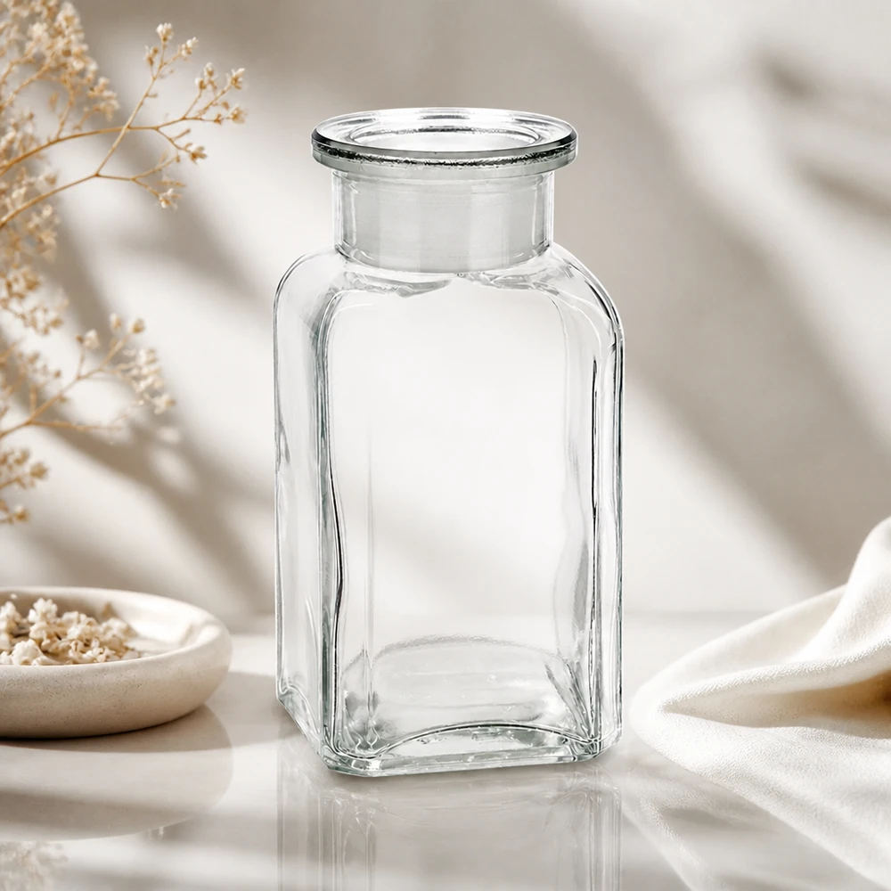 Botella cuadrada transparente y vacía de 100 ml con cuello ancho, sobre un elegante fondo de tela blanca y flores secas