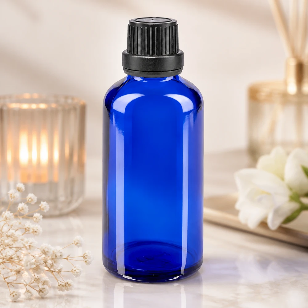 Frasco de cristal azul cobalto de 50 ml con tapón de rosca negro y gotero, junto a una vela y unas flores