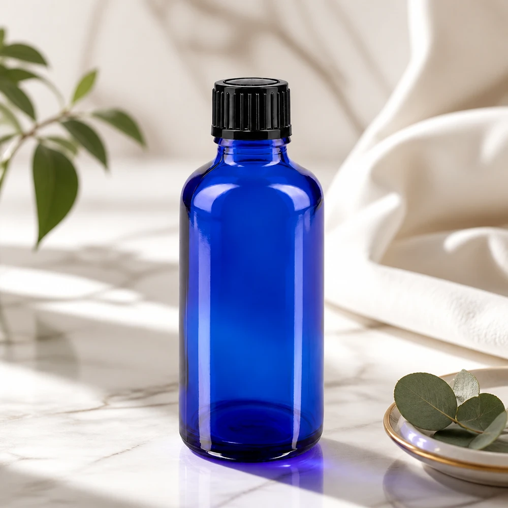 Botella de cristal azul de 50 ml con tapón negro sobre mármol, junto a hojas de eucalipto