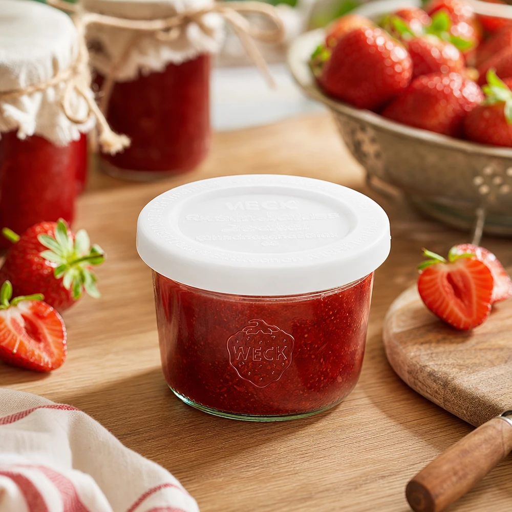 Vasos WECK RR60 de 80 ml llenos de mermelada de fresa sobre una tabla de madera rodeados de fresas frescas y tarros de mermelada