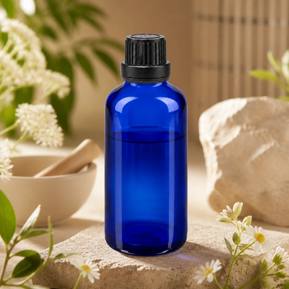 Frasco de cristal azul cobalto de 50 ml con tapón de rosca negro, sobre una piedra natural con flores de manzanilla y un mortero