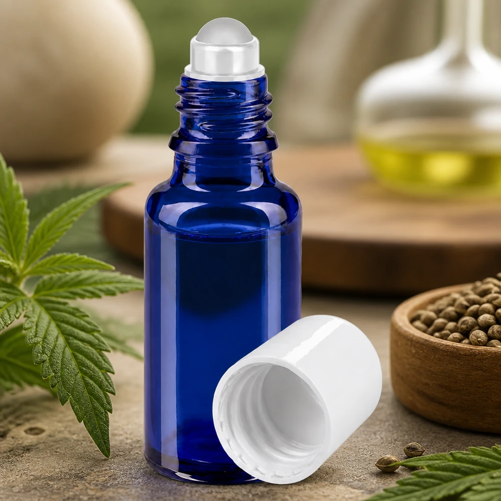 Frasco de cristal azul de 20 ml con aplicador de bola y tapón blanco, rodeado de hojas y semillas de cáñamo
