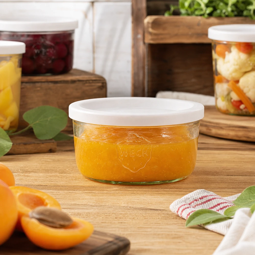 Vaso de 165ml con tapa de frescura WECK RR80 Tarro WECK de 165 ml lleno de mermelada de naranja en un entorno de cocina natural con tarros de conserva al fondo.