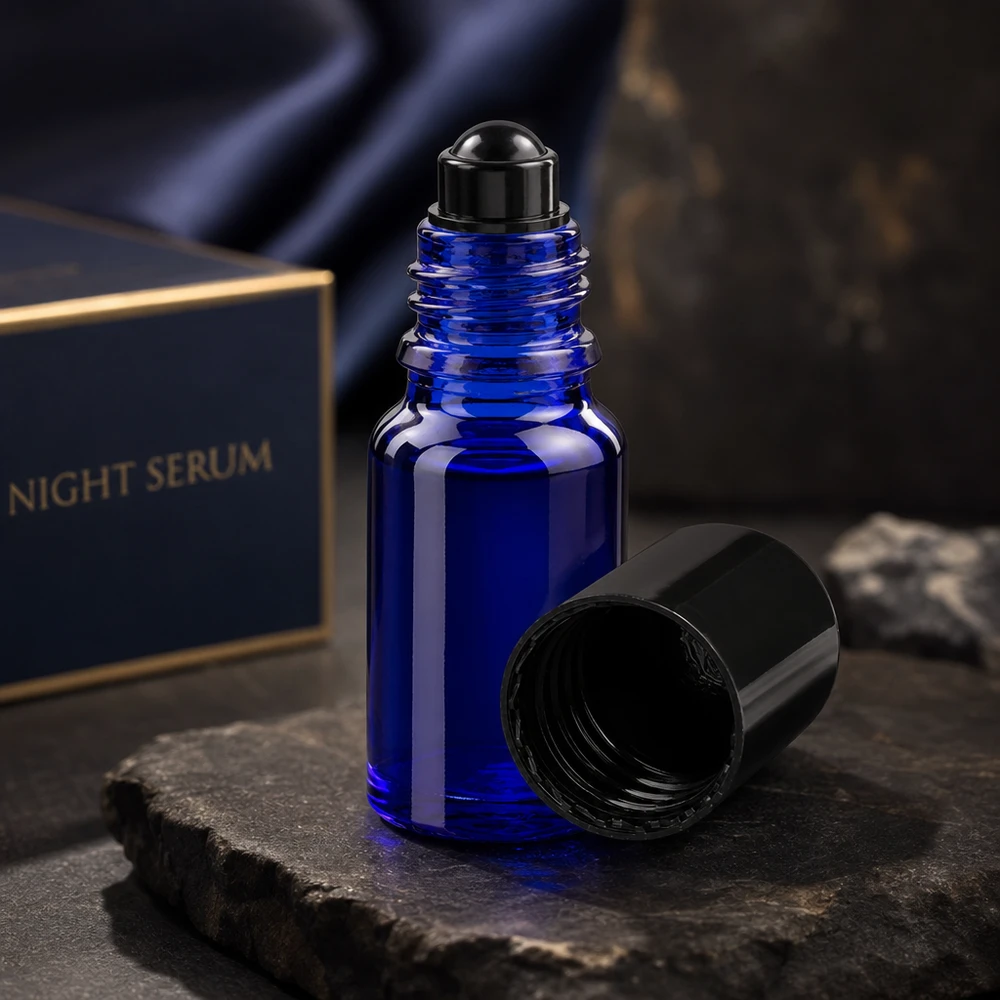 Botella de farmacia azul 10ml tapón roll-on negro Frasco roll-on ApoGlas azul oscuro de 10 ml con la tapa negra abierta, sobre una piedra de pizarra, delante de la caja del sérum de noche