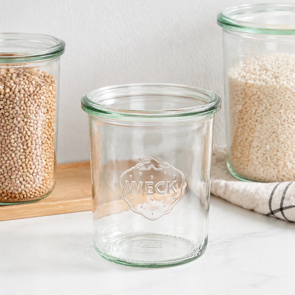 Tarro de conserva de 160ml WECK RR60 WECK 160ml RR60 mason jar con borde verde y logo, mason jar vacío junto a tarros de almacenaje llenos sobre tabla de madera