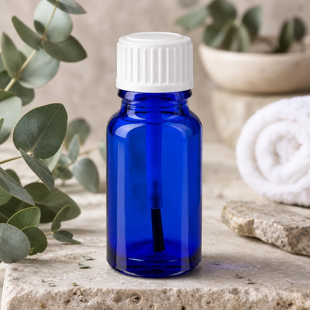 Botella de farmacia azul 10ml tapón rosca cepillo blanco Frasco de cristal azul de 10 ml con un aplicador de pincel blanco junto a unas ramas de eucalipto sobre piedra natural