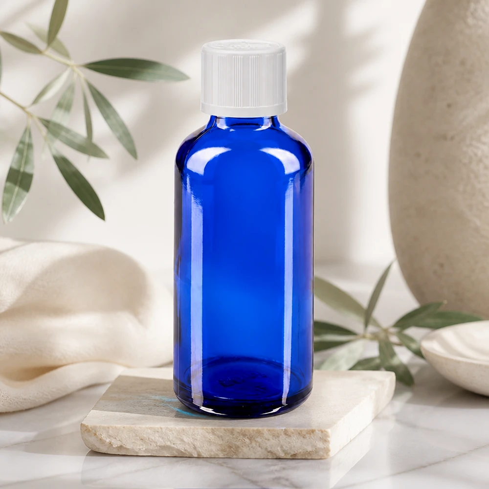 Botella de cristal azul de 50 ml con tapón de rosca blanco sobre una losa de mármol en un ambiente natural y luminoso